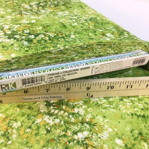 May include: A roll of fabric with a meadow-themed print. The fabric features a green background with white and orange floral accents. The fabric is labeled "FUSIONS & COLLECTION GARDEN" and is 100% cotton. A wooden ruler is placed on top of the fabric.