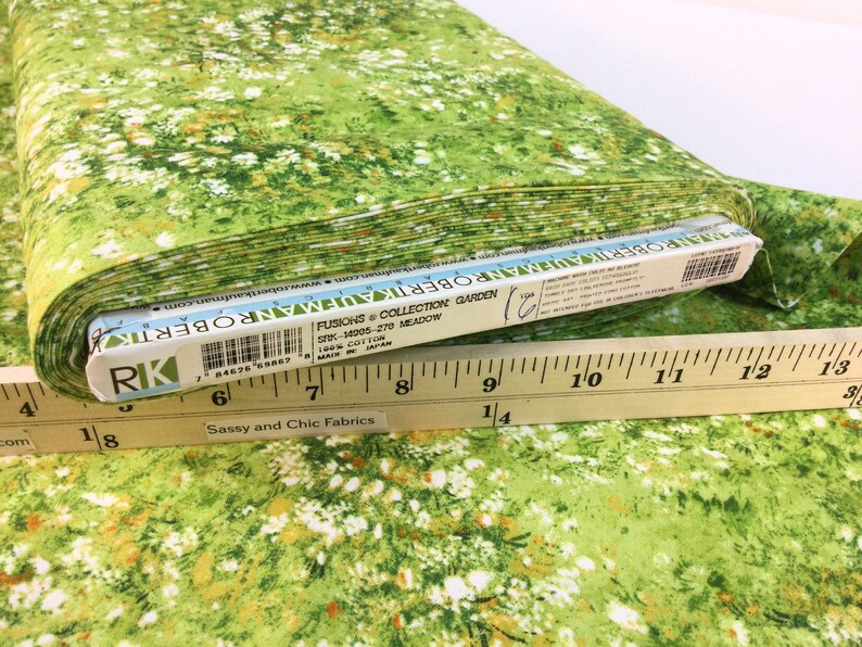 May include: A roll of fabric with a meadow-themed print. The fabric features a green background with white and orange floral accents. The fabric is labeled "FUSIONS & COLLECTION GARDEN" and is 100% cotton. A wooden ruler is placed on top of the fabric.