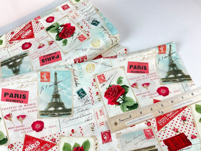 Może przedstawiać: Tkanina z motywem vintage poczt&oacute;wki z Wieżą Eiffla, czerwonymi r&oacute;żami i znaczkami pocztowymi. Tkanina ma kremowe tło z czerwonymi i niebieskimi akcentami. Na tkaninie umieszczono linijkę.