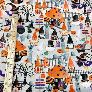 May include: Halloween-themed fabric featuring gnomes, pumpkins, tombstones, and witch hats on a light gray background. The fabric includes orange, black, purple, and white elements. The design incorporates a ruler for scale, with measurements in inches.
