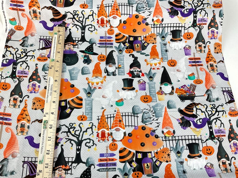 May include: Halloween-themed fabric featuring gnomes, pumpkins, tombstones, and witch hats on a light gray background. The fabric includes orange, black, purple, and white elements. The design incorporates a ruler for scale, with measurements in inches.