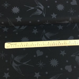 May include: Black fabric featuring a repeating pattern of grey swallows, stars, hearts, and dots. A ruler is placed across the fabric, showing inches. The fabric is folded over, revealing more of the pattern.