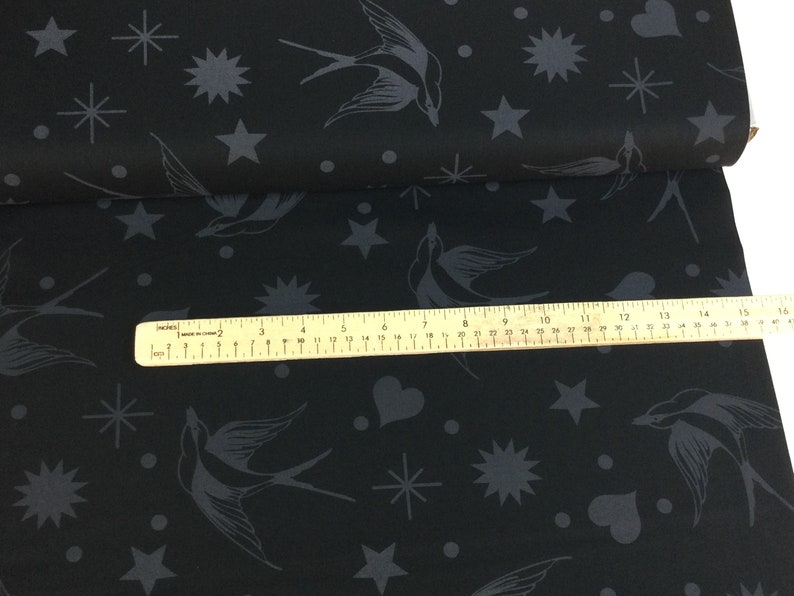May include: Black fabric featuring a repeating pattern of grey swallows, stars, hearts, and dots. A ruler is placed across the fabric, showing inches. The fabric is folded over, revealing more of the pattern.