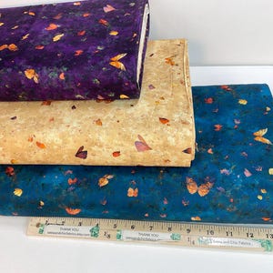 May include: Three stacks of fabric with butterfly patterns. The top fabric is a deep purple with orange and yellow butterflies. The middle fabric is tan with orange and purple butterflies. The bottom fabric is teal with orange and purple butterflies. A ruler is at the bottom.