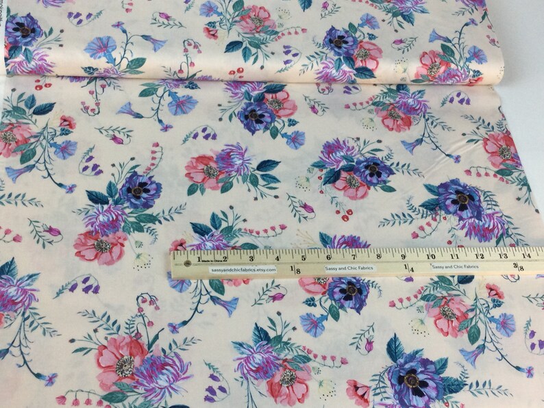 May include: Cream fabric with a floral design of pink, purple, and blue flowers. The fabric has a ruler along the bottom edge. The flowers are arranged in clusters with green leaves and stems.