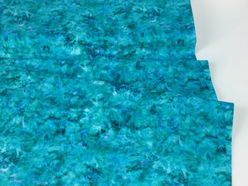May include: A close-up of a fabric with a watercolor-like pattern in shades of teal, turquoise, and blue. The fabric has a textured appearance, with areas of darker and lighter hues creating a sense of depth and movement. The fabric is folded.