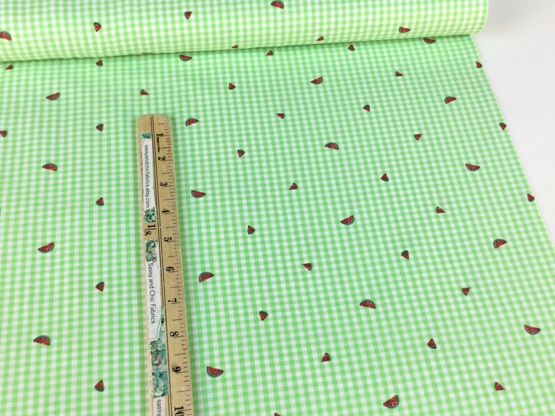 K&ouml;nnte beinhalten: Gr&uuml;n-wei&szlig;es Vichy-Stoff mit einem sich wiederholenden Muster aus Wassermelonenscheiben. Ein Holzlineal mit Zentimetermarkierungen liegt auf dem Stoff. Der Stoff ist im Hintergrund aufgerollt.