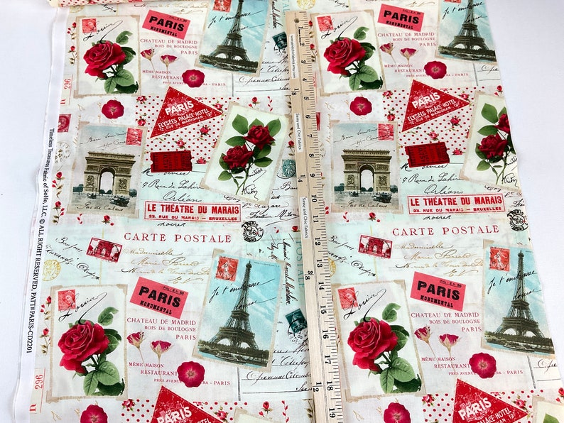 Może przedstawiać: Tkanina o tematyce paryskiej z czerwonymi r&oacute;żami, ilustracjami wieży Eiffla i wzorami poczt&oacute;wek. Tkanina zawiera tekst taki jak "Paris" i "Carte Postale". Drewniana linijka jest umieszczona na tkaninie.