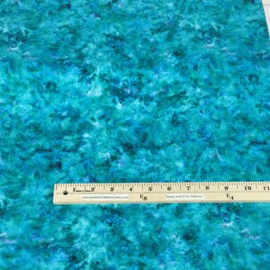 May include: A piece of fabric with a teal and blue watercolor-like pattern. A wooden ruler with inch measurements is placed on the fabric. The fabric has a mottled appearance, with darker blue areas interspersed throughout the lighter teal.