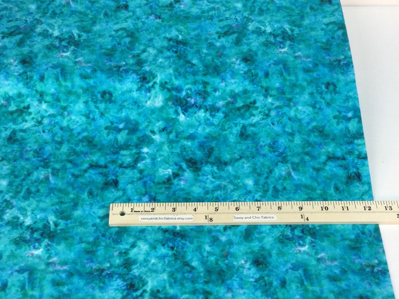 May include: A piece of fabric with a teal and blue watercolor-like pattern. A wooden ruler with inch measurements is placed on the fabric. The fabric has a mottled appearance, with darker blue areas interspersed throughout the lighter teal.