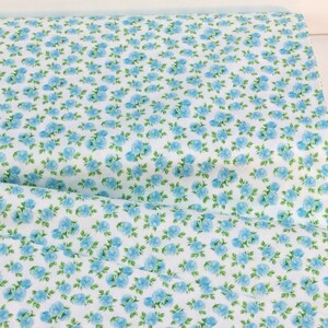 May include: White fabric with a repeating pattern of small, light blue roses and green leaves. The floral design covers the entire surface of the fabric, creating a dense, all-over pattern. The fabric is folded, showing the pattern's continuity.