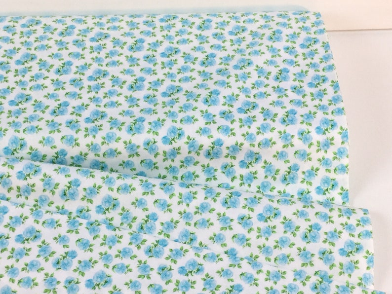 May include: White fabric with a repeating pattern of small, light blue roses and green leaves. The floral design covers the entire surface of the fabric, creating a dense, all-over pattern. The fabric is folded, showing the pattern's continuity.