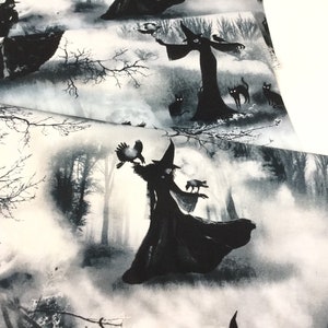 May include: Fabric with a black and white witch-themed print. The design features silhouetted witches, black cats, crows, and bare trees against a misty forest backdrop. The fabric is folded, showing the pattern's repeat.