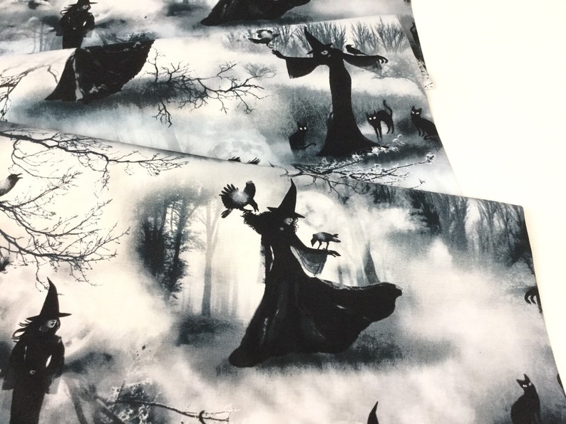 May include: Fabric with a black and white witch-themed print. The design features silhouetted witches, black cats, crows, and bare trees against a misty forest backdrop. The fabric is folded, showing the pattern's repeat.