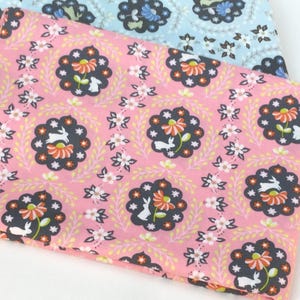 May include: Two folded fabric pieces with floral and bunny patterns. The top fabric is pink with a repeating pattern of dark gray floral medallions, white bunnies, and orange flowers. The bottom fabric is light blue with a similar floral design.