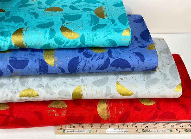 May include: A stack of colorful fabric bolts featuring a repeating pattern of gold half-circles on a variety of backgrounds. Colors include turquoise, blue, light blue, white, and red. A wooden ruler is at the bottom.