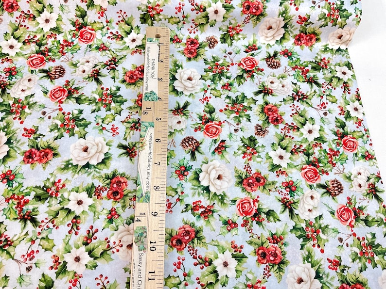 K&ouml;nnte beinhalten: Blumenstoff mit hellblauem Hintergrund, mit roten Rosen, wei&szlig;en Blumen, gr&uuml;nen Bl&auml;ttern und roten Beeren. Ein Holzlineal ist vertikal auf dem Stoff platziert. Auf dem Lineal steht "THANK YOU" und "Sassy and Chic Fabrics".