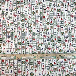 May include: White fabric with a repeating pattern of Mahjong tiles in red, green, and brown. The tiles feature Chinese characters, flowers, and other symbols. A wooden ruler with inch markings is placed across the fabric.