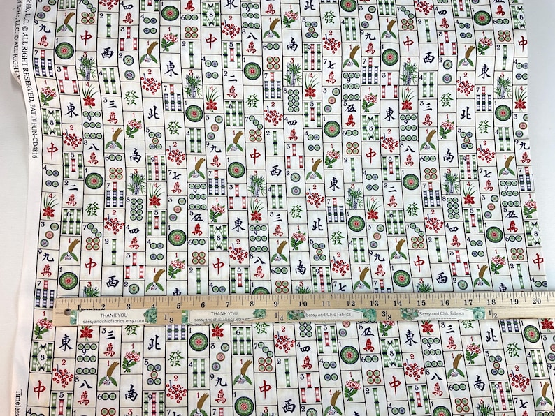 May include: White fabric with a repeating pattern of Mahjong tiles in red, green, and brown. The tiles feature Chinese characters, flowers, and other symbols. A wooden ruler with inch markings is placed across the fabric.