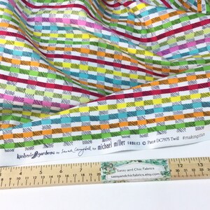 May include: Colorful fabric with a geometric pattern of horizontal and vertical lines in various colors, including red, green, blue, and orange. The fabric has the text "Kashmir Gardens" and "Michael Miller Fabrics". A ruler shows measurements in inches.