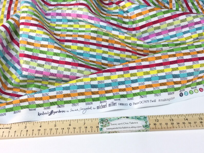 May include: Colorful fabric with a geometric pattern of horizontal and vertical lines in various colors, including red, green, blue, and orange. The fabric has the text "Kashmir Gardens" and "Michael Miller Fabrics". A ruler shows measurements in inches.