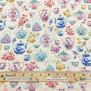 May include: Cream-colored fabric patterned with various teapots and teacups in shades of blue, pink, and green, adorned with floral arrangements. A wooden ruler with inch markings is at the bottom.