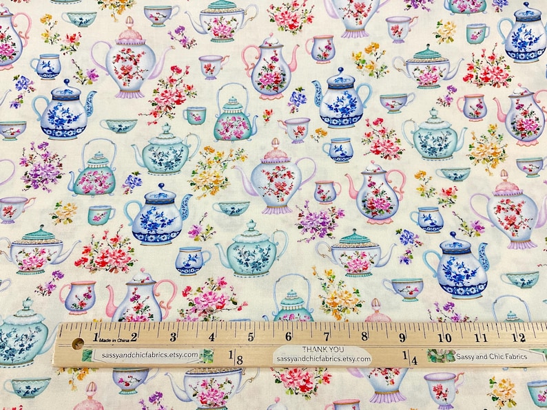 May include: Cream-colored fabric patterned with various teapots and teacups in shades of blue, pink, and green, adorned with floral arrangements. A wooden ruler with inch markings is at the bottom.
