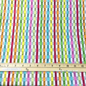 May include: Colorful fabric with a repeating pattern of vertical stripes and rectangular blocks in shades of red, pink, orange, yellow, green, and blue. A ruler is placed across the fabric, showing measurements in inches.