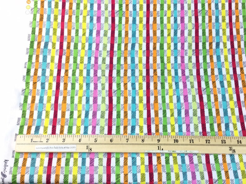 May include: Colorful fabric with a repeating pattern of vertical stripes and rectangular blocks in shades of red, pink, orange, yellow, green, and blue. A ruler is placed across the fabric, showing measurements in inches.
