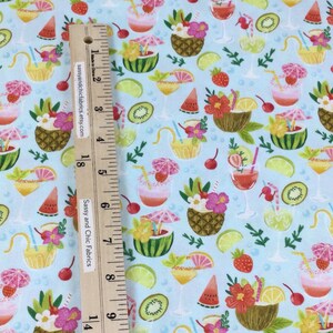 May include: Light blue fabric with a tropical cocktail pattern. The design features various cocktails, including those in pineapple, coconut, and watermelon, along with fruit slices and flowers. A wooden ruler with text is placed on the fabric.