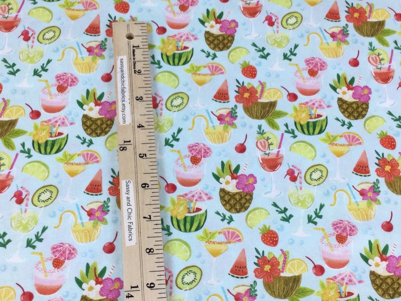 May include: Light blue fabric with a tropical cocktail pattern. The design features various cocktails, including those in pineapple, coconut, and watermelon, along with fruit slices and flowers. A wooden ruler with text is placed on the fabric.