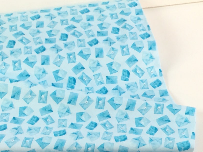 May include: Light blue fabric with a repeating pattern of watercolor-style envelopes. The envelopes are in varying shades of blue and have a button closure design. The fabric is likely cotton or a similar material, suitable for quilting or crafting projects.