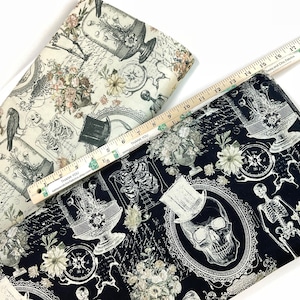 May include: Two fabric pieces with a gothic theme. One is cream-colored with skeletons, flowers, and vintage illustrations. The other is black with similar designs, including a skull in a top hat. A wooden ruler is placed across the fabrics.
