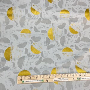 May include: Fabric with a repeating pattern of gold and gray semi-circles and brushstrokes on a light gray background. A wooden ruler with inch markings is placed across the fabric. The fabric is from the "Wild Rose Collection".