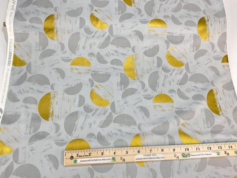 May include: Fabric with a repeating pattern of gold and gray semi-circles and brushstrokes on a light gray background. A wooden ruler with inch markings is placed across the fabric. The fabric is from the "Wild Rose Collection".