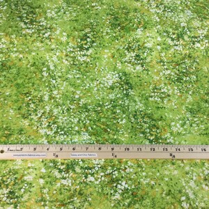 May include: Green fabric with a pattern of white and brown flowers. The fabric has a grassy appearance, with a ruler placed across the bottom. The fabric is likely used for sewing or crafting projects.