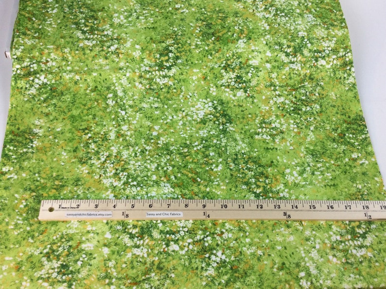 May include: Green fabric with a pattern of white and brown flowers. The fabric has a grassy appearance, with a ruler placed across the bottom. The fabric is likely used for sewing or crafting projects.