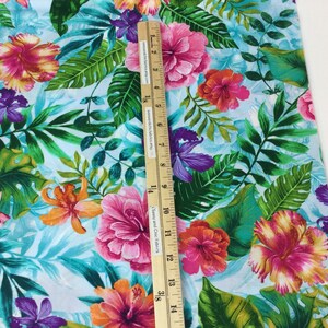 May include: Vibrant fabric with a tropical floral pattern. The design features large pink, orange, and purple hibiscus flowers and green leaves on a light blue background. A wooden ruler is placed on the fabric, showing measurements in inches.