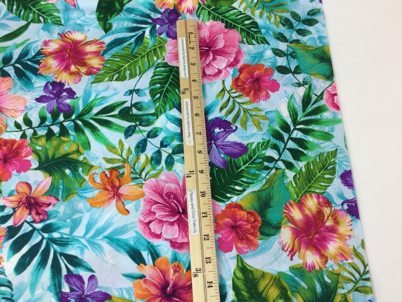 May include: Vibrant fabric with a tropical floral pattern. The design features large pink, orange, and purple hibiscus flowers and green leaves on a light blue background. A wooden ruler is placed on the fabric, showing measurements in inches.
