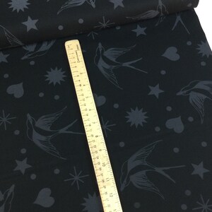 May include: Black fabric featuring a pattern of swallows, stars, hearts, and dots in dark grey. A wooden ruler is placed diagonally across the fabric, showing centimetre measurements.