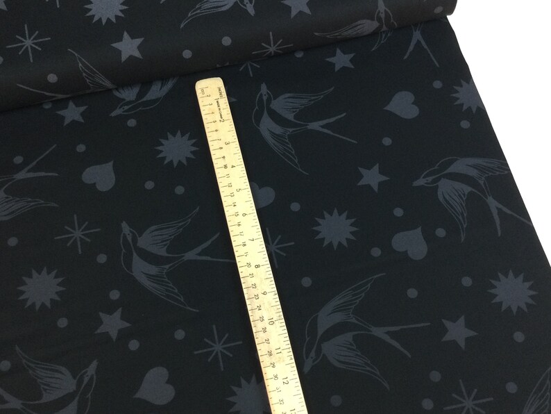 May include: Black fabric featuring a pattern of swallows, stars, hearts, and dots in dark grey. A wooden ruler is placed diagonally across the fabric, showing centimetre measurements.