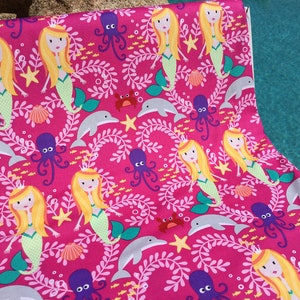 May include: Hot pink fabric with a repeating pattern of mermaids, dolphins, octopuses, starfish, and seashells. The mermaids have blonde hair and green tails. The fabric is likely for crafting or sewing projects.
