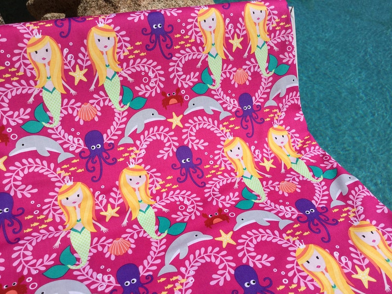 May include: Hot pink fabric with a repeating pattern of mermaids, dolphins, octopuses, starfish, and seashells. The mermaids have blonde hair and green tails. The fabric is likely for crafting or sewing projects.