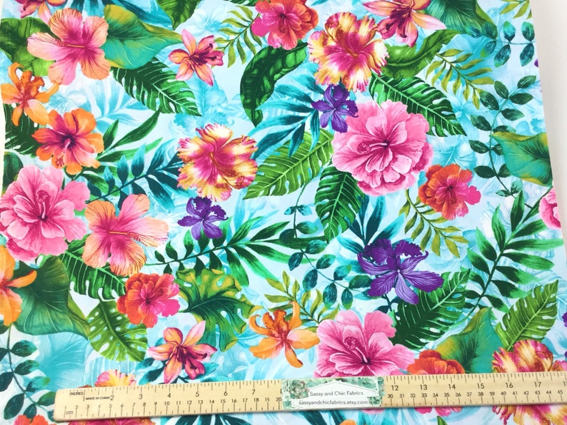 May include: Vibrant fabric featuring a tropical floral pattern. Large pink, orange, and yellow hibiscus flowers are interspersed with green leaves and purple orchids against a light blue background. A ruler is visible along the bottom edge.