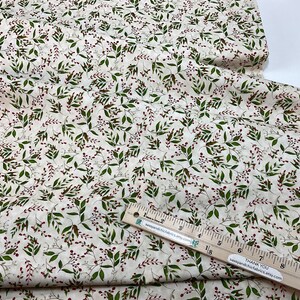Puede incluir: Tela de color crema con un patr&oacute;n repetido de hojas verdes y peque&ntilde;as bayas rojas. La tela est&aacute; doblada y parece ser de algod&oacute;n o lino. Se ve una regla de madera con marcas en pulgadas, que indica las dimensiones de la tela.
