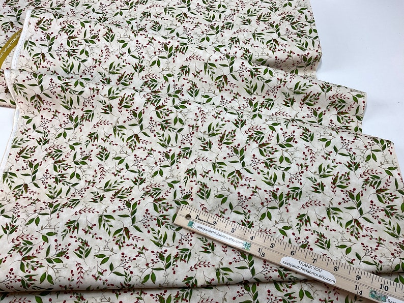 Puede incluir: Tela de color crema con un patr&oacute;n repetido de hojas verdes y peque&ntilde;as bayas rojas. La tela est&aacute; doblada y parece ser de algod&oacute;n o lino. Se ve una regla de madera con marcas en pulgadas, que indica las dimensiones de la tela.