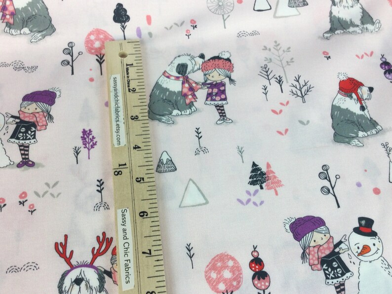May include: Light pink fabric with a winter scene featuring cartoon dogs, children, trees, and snowmen. The dogs wear hats and scarves. The children wear hats, scarves, and dresses. The fabric has a ruler with the text "Sassy and Chic Fabrics etsy.com".