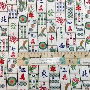 May include: Mahjong tile fabric with a cream background. The fabric features a repeating pattern of Mahjong tiles with Chinese characters, bamboo, flowers, and circles in red, green, and blue. A wooden ruler is placed on top.