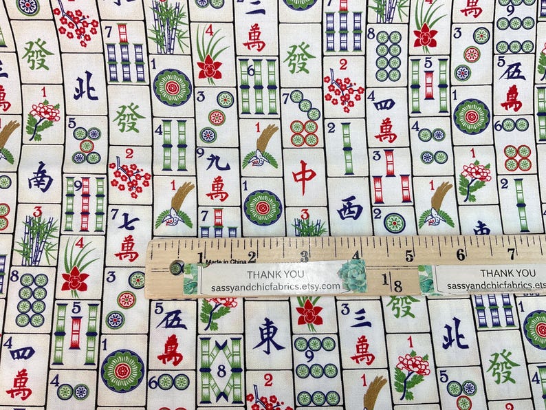 May include: Mahjong tile fabric with a cream background. The fabric features a repeating pattern of Mahjong tiles with Chinese characters, bamboo, flowers, and circles in red, green, and blue. A wooden ruler is placed on top.