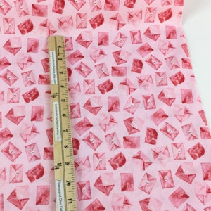 May include: Pink fabric with a repeating pattern of watercolor-style red envelopes. A wooden ruler with inch measurements is placed on the fabric. The fabric is likely for crafting or sewing projects.